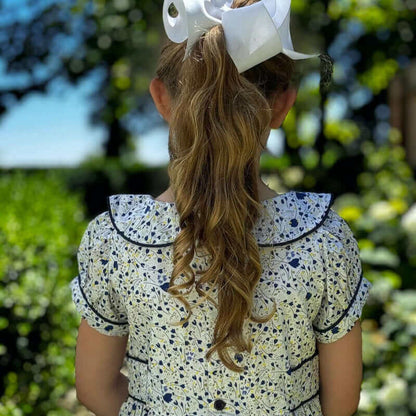 Girl wearing Jane Ruffle Dress with piped Peter Pan collar and floral print, standing in garden with white hair bow.