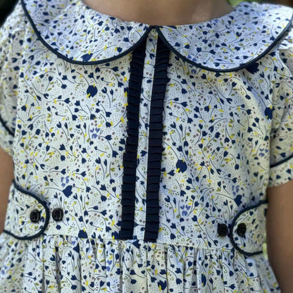 Crisp blue floral print Jane Ruffle Dress with a piped Peter Pan collar, ruffled placard, capped sleeves, navy buttons, and front pockets