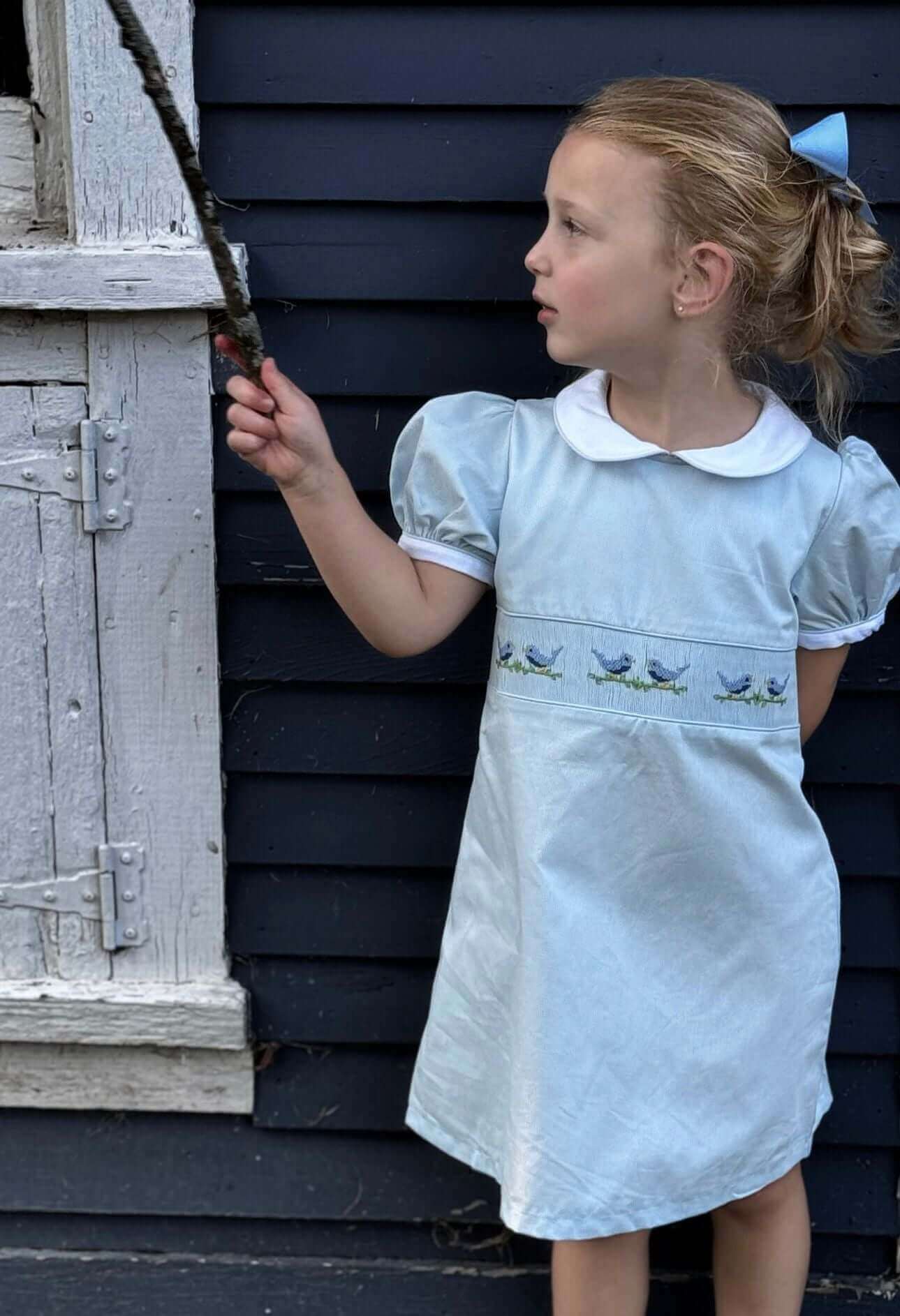 Young girl in Smocked Sky Lovebird Dress standing by a vintage wooden window.