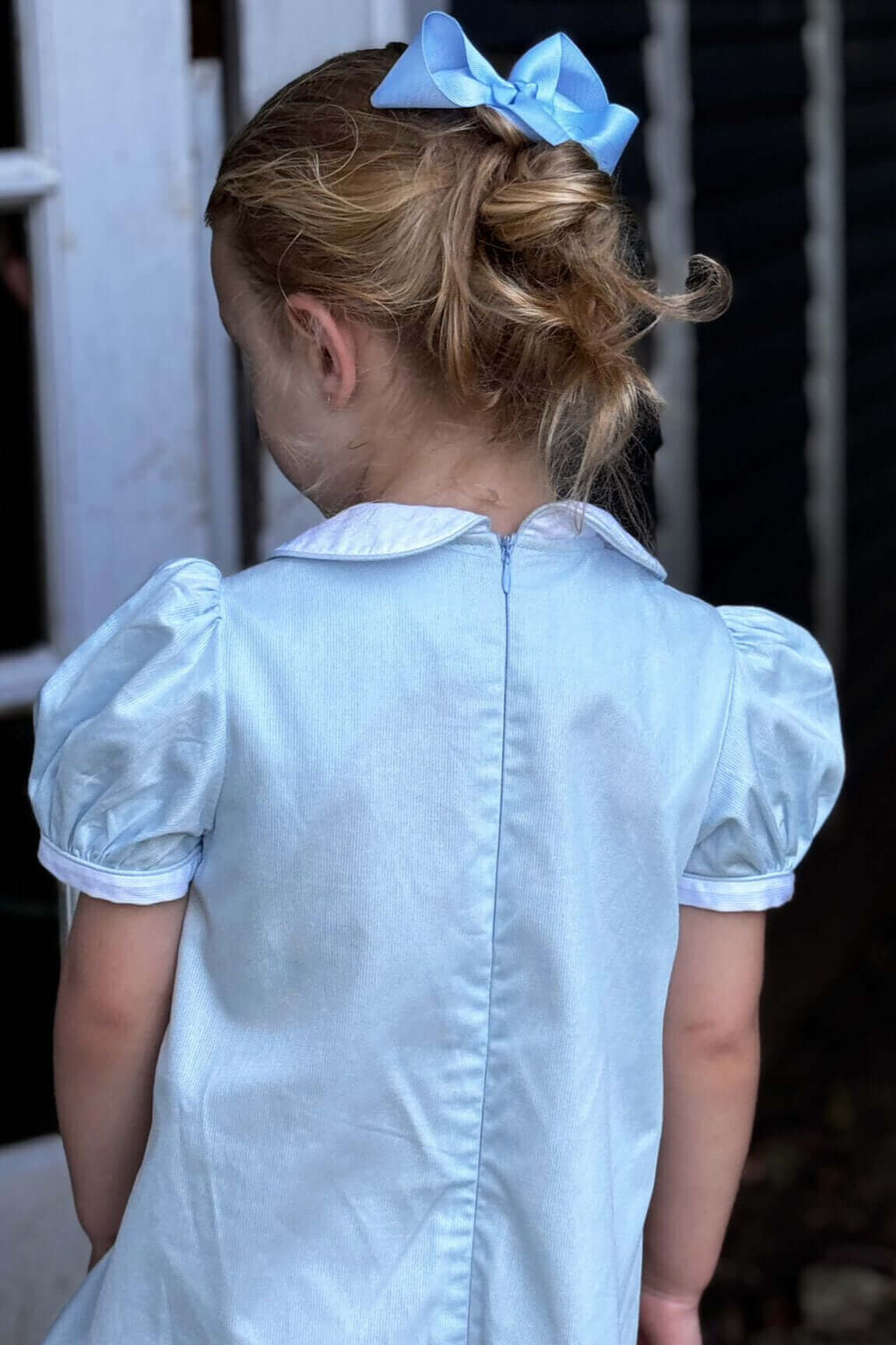 Young girl in a Smocked Sky Lovebird Dress with a blue bow, showcasing the back view of the hand-smocked elegant dress.