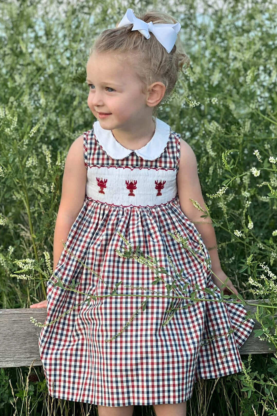 Little girl in a smocked lobster dress with red, white, and blue checks, standing in a field, perfect for summer fun and family photos.