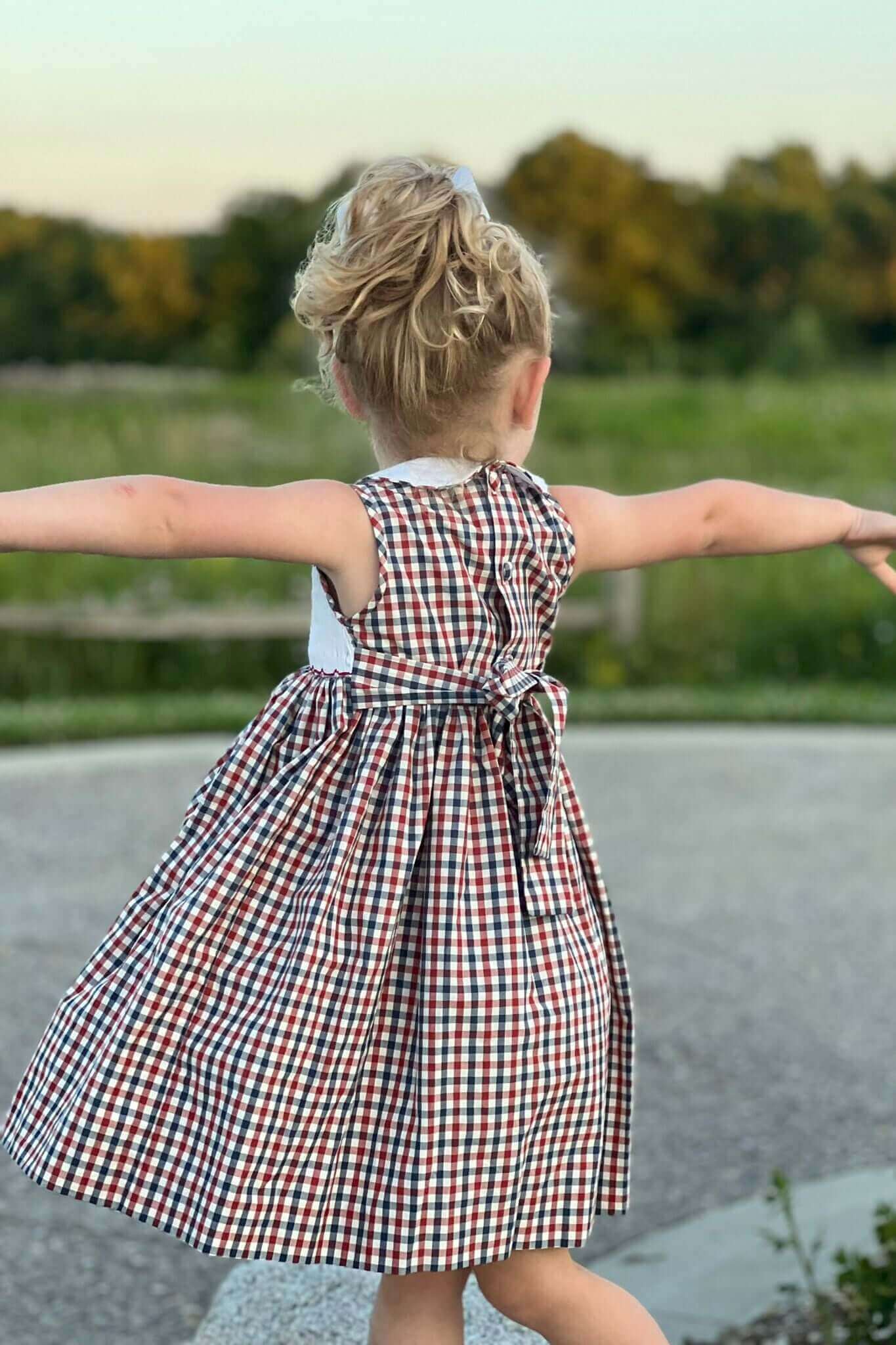 Young girl twirls in a Smocked Lobster Dress with red, white, and blue checks, enjoying a sunny day outdoors.