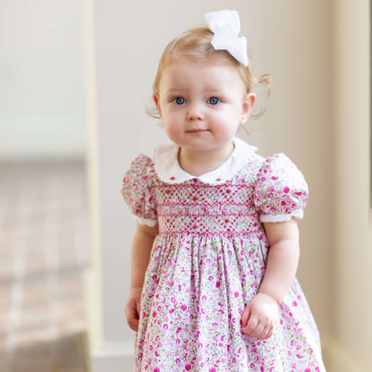 Adorable toddler in a hand-smocked floral dress with a white collar and bow, showcasing intricate craftsmanship.