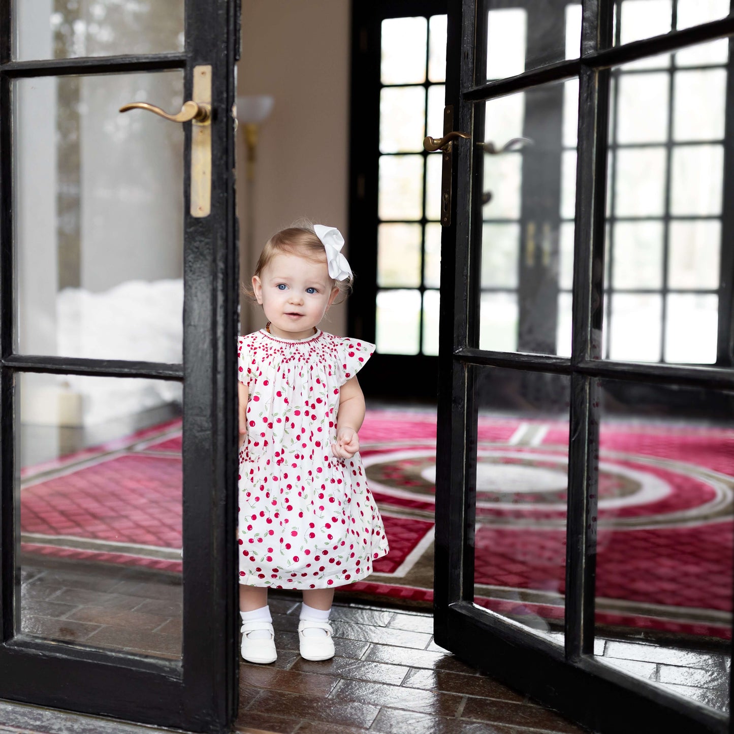 Adorable baby girl wearing a Smocked Cherry Bishop Dress with a bow, standing at a doorway.