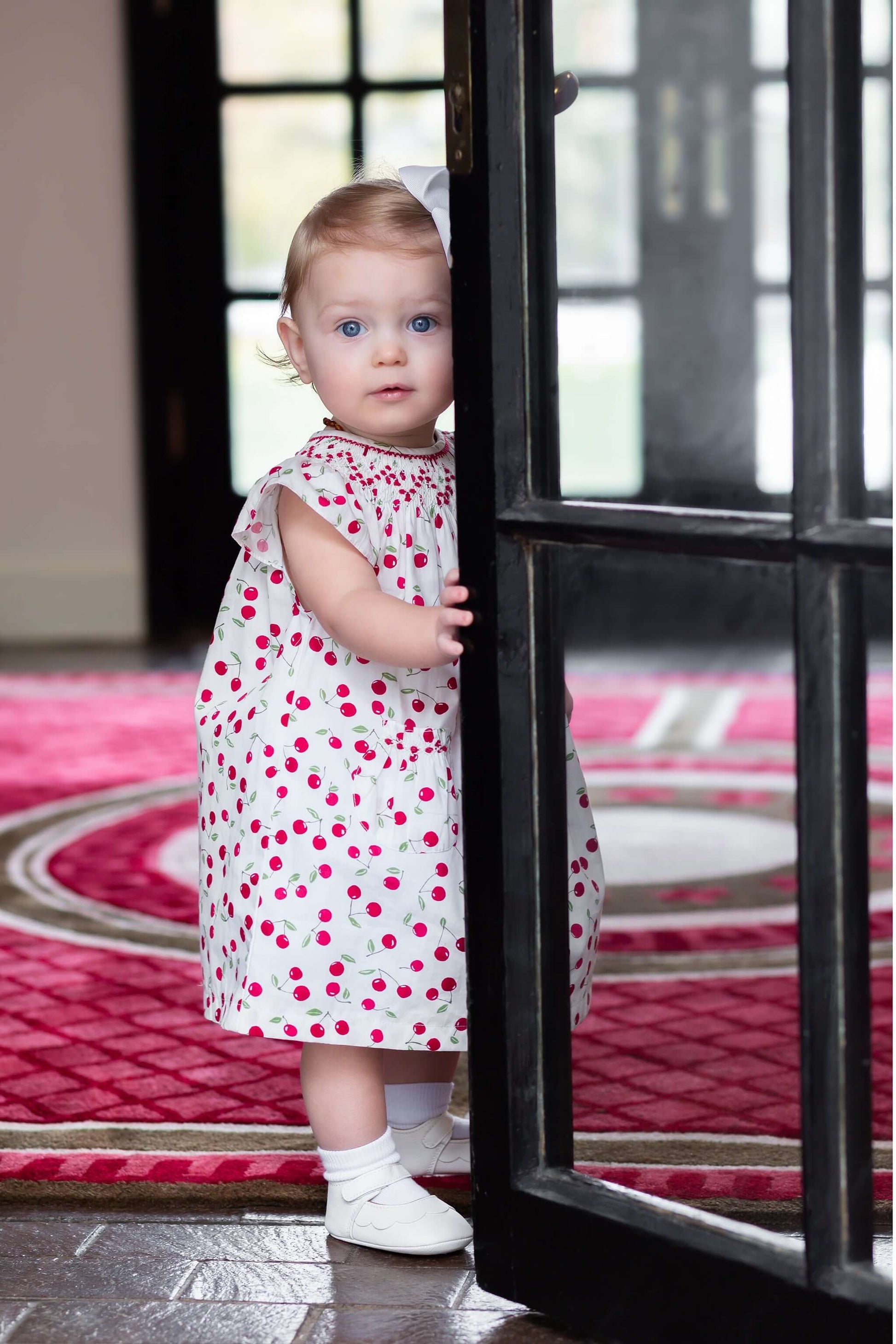 Infant girl peeking through a door wearing a smocked cherry bishop dress, featuring a playful cherry print.