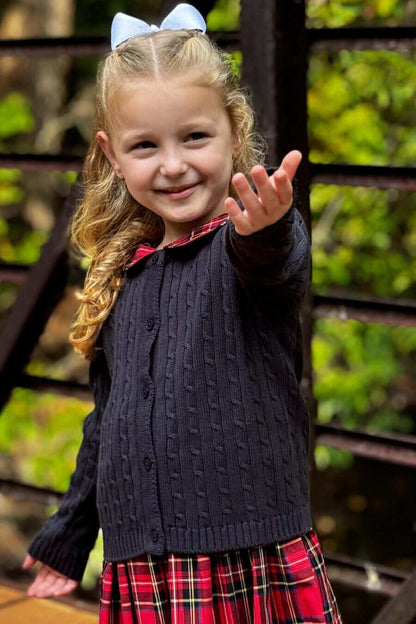 Little girl wears a navy cotton cable knit cardigan with a red plaid skirt, reaching out with a smile.