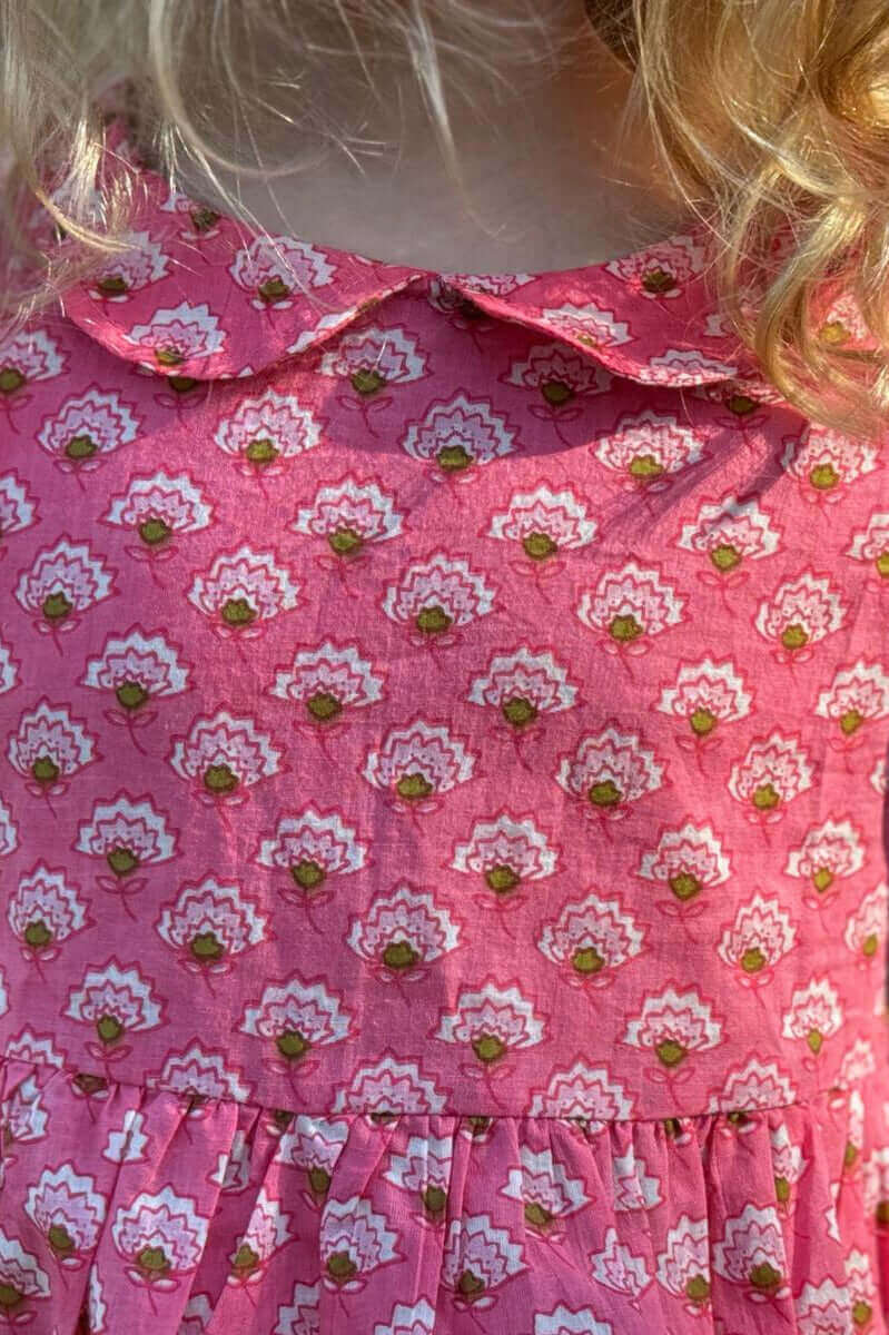 Child wearing the Pinkberry Classic Dress with floral print, featuring a Peter Pan collar and smocked design in pink fabric.