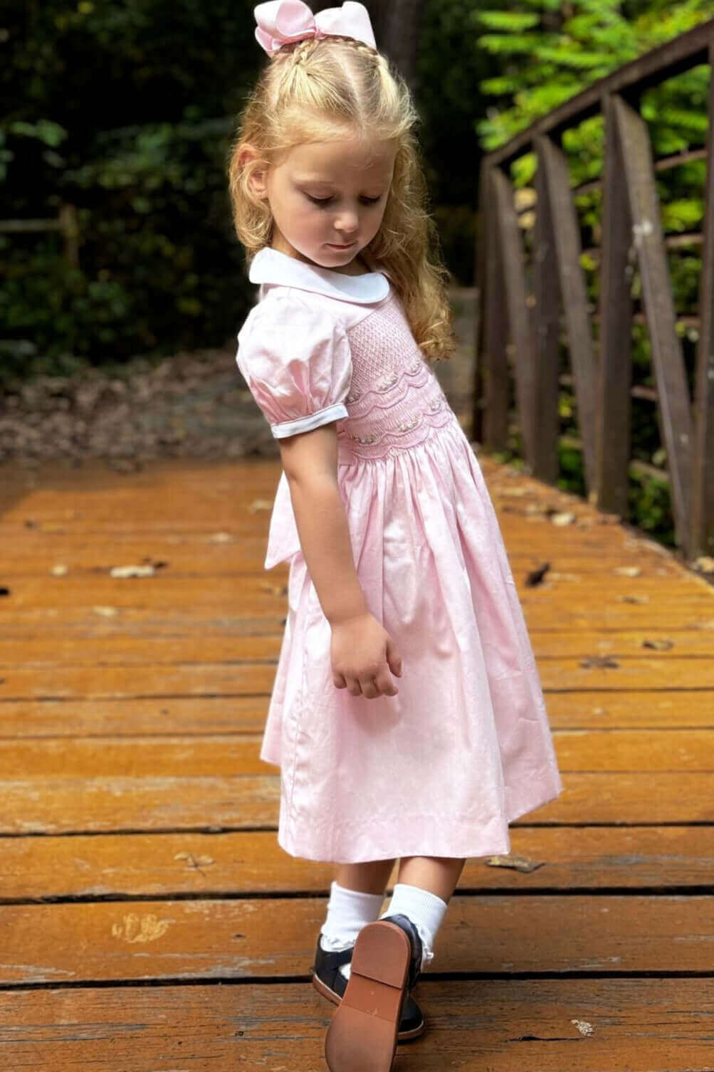 Little girl in a Smocked Pink Garland Dress with covered buttons and attached belt, standing on a wooden bridge looking down.