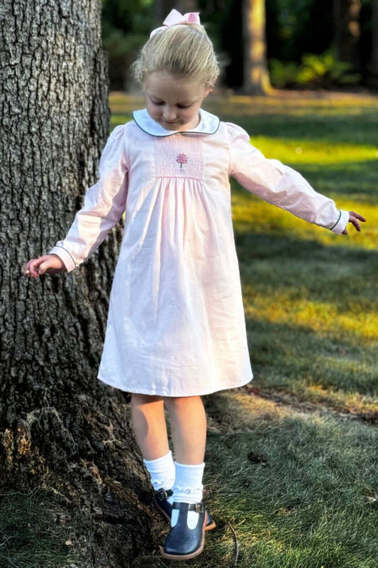 Little girl in a Smocked Pink Topiary Dress with hand-smocked details, posed by a tree in a sunlit park.