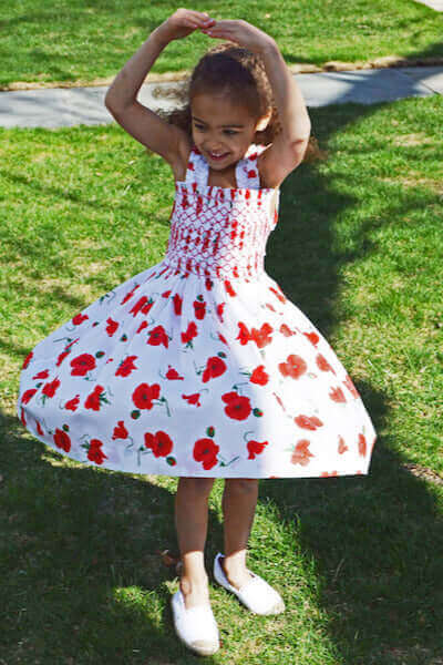 Child twirling in vibrant smocked poppy sundress with intricate lattice embroidery, showcasing summer style and craftsmanship.