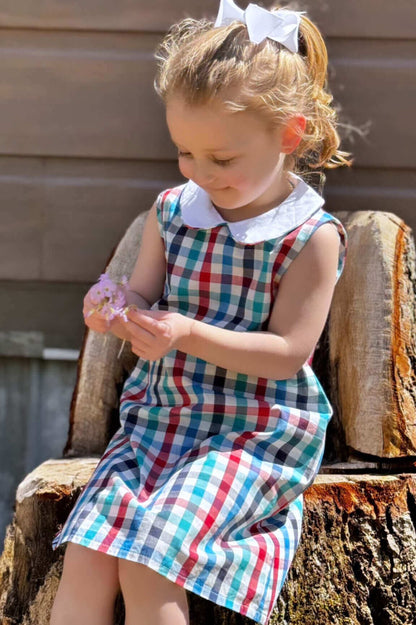 Child wearing a preppy plaid dress with a Peter Pan collar, sitting outdoors on a wooden log, holding a flower.