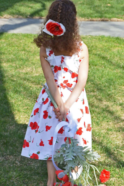 Little girl wearing a hand-smocked poppy sundress with vibrant red flowers, enjoying a sunny day in the garden.