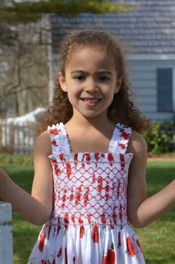 Young girl wearing a vibrant Smocked Poppy Sundress with delicate lattice embroidery, smiling in a garden setting.