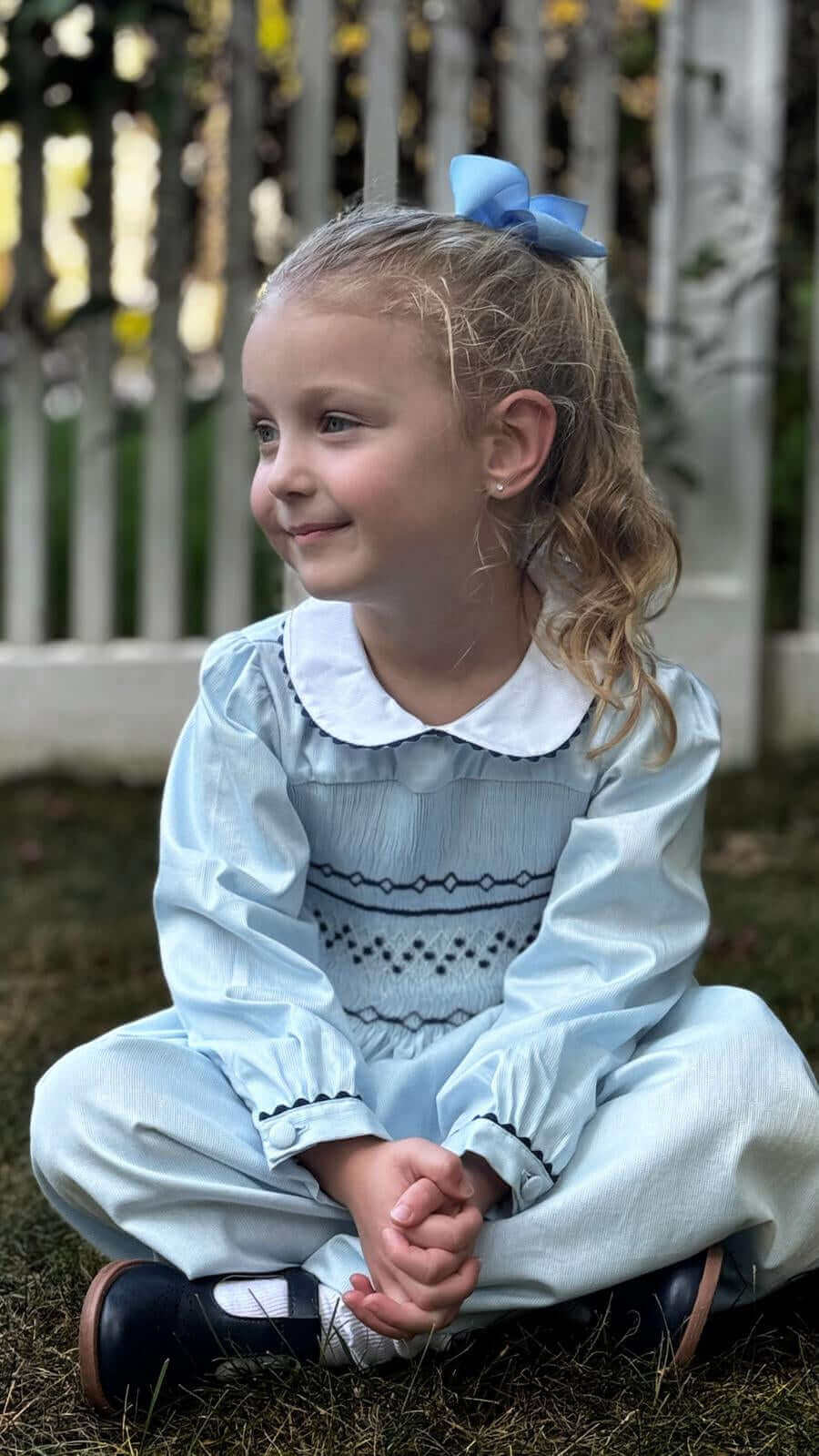 A little girl sits on grass wearing a Smocked Sky Blue Geo Dress, smiling softly, with a matching blue bow and a white picket fence behind her.