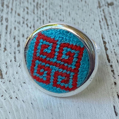 Blue and red Greek key needlepoint ring with silver adjustable band on a rustic wooden background.