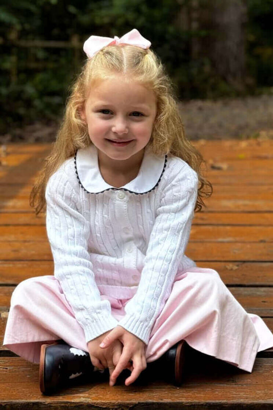 Little girl wearing a white cotton cable knit cardigan, pink dress, and a matching pink bow sitting on a wooden pathway