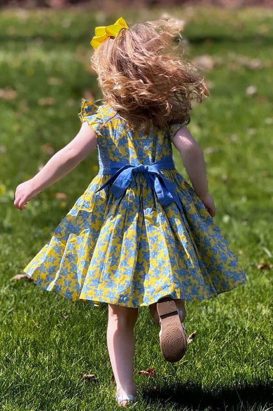 Little girl running in a yellow and blue floral Forget Me Not dress with a cornflower belt, enjoying a sunny day in the park.