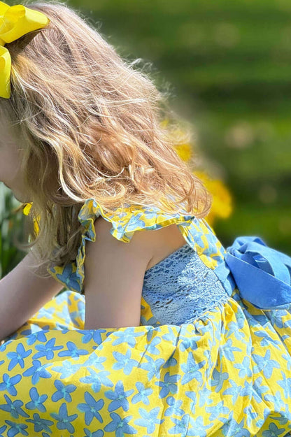 Child in Forget Me Not yellow and blue cotton floral dress with cornflower belt, enjoying a sunny day outdoors.