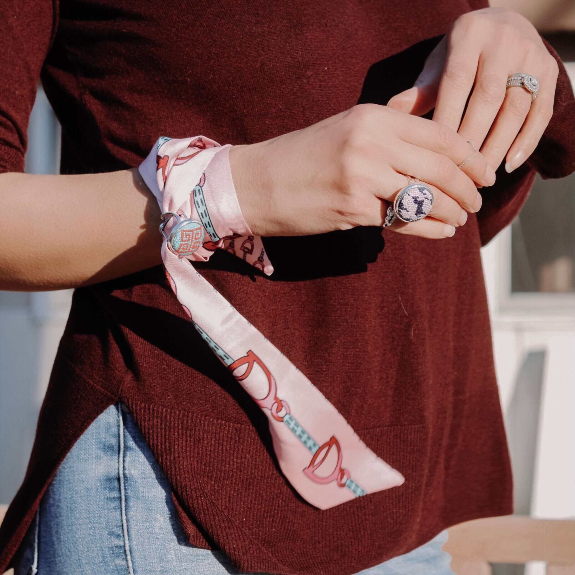 Woman wearing a pink scarf tied around her wrist, showcasing a blue and red Greek Key needlepoint ring on her finger.