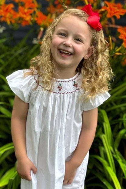 Smocked Cherry Bishop Dress (6) worn by a smiling girl with curly hair and a red bow, surrounded by flowers.