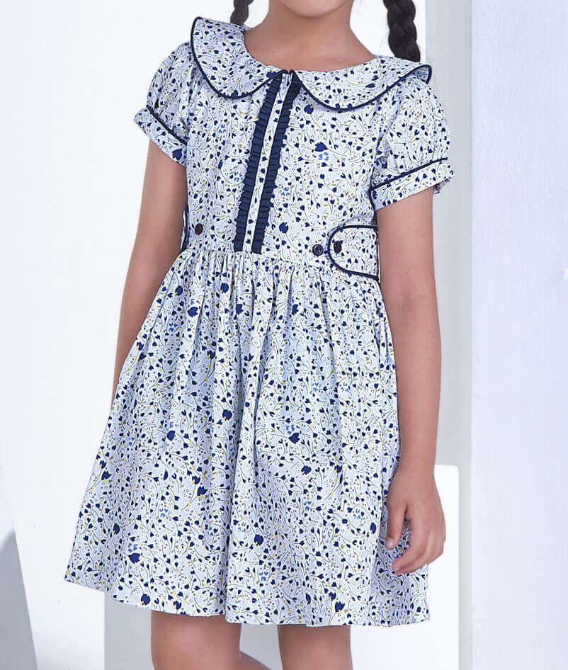 Girl wearing the Jane Ruffle Dress in crisp blue floral, featuring a piped Peter Pan collar, ruffled placket, capped sleeves, and navy buttons.